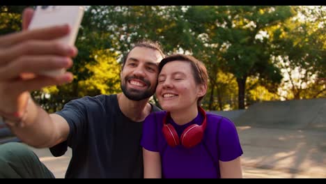 A-guy-in-a-gray-t-shirt-and-a-girl-with-a-short-haircut-in-a-purple-top-with-red-headphones-take-a-selfie-using-a-white-phone-in-a-park-in-summer