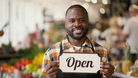 Portrait-of-a-Black-skinned-man-in-a-plaid-shirt-and-black-apron-holding-a-sign-with-the-sign-Open-in-a-supermarket