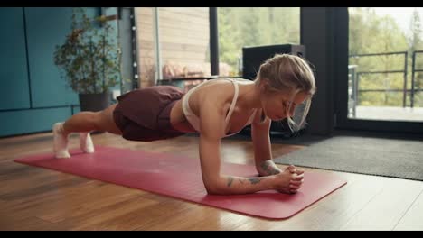 A-blonde-girl-with-tattoos-in-a-white-top-does-a-plank-exercise-on-a-special-mat-in-an-industrial-house-overlooking-a-green-forest