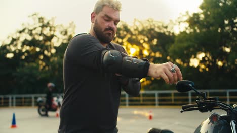 A-man-biker-and-strong-build-with-a-thick-beard-puts-on-elbow-pads-for-protection-while-eating-on-a-motorcycle