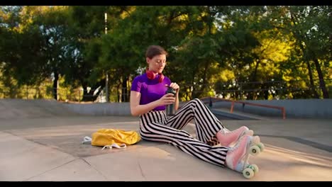 a-girl-with-a-short-haircut-with-a-purple-top-and-striped-pants-in-pink-roller-skates-sits-in-a-skate-park-on-a-concrete-floor-and-drinks-water-from-a-special-bottle