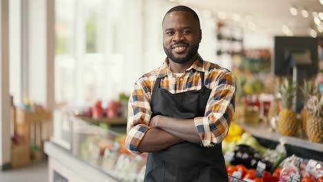 Portrait-of-a-Black-skinned-man-in-a-plaid-shirt-and-black-apron-folded-his-arms-over-his-chest-and-looks-at-the-camera-in-a-supermarket