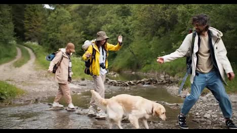 The-guy-helps-two-girls-to-cross-a-mountain-river-on-special-stones,-he-gives-him-a-hand-so-that-they-grab-it-and-cross-to-the-other-side.-People-in-hiking-clothes-ourdoor