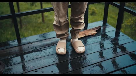 Close-up-shot:-a-person-in-slippers-stands-on-a-floor-of-dark-boards-during-the-rain-outside