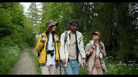 A-guy-and-two-girls-in-special-hiking-clothes-are-walking-along-a-path-along-a-green-forest.-Chatting-while-walking-through-the-woods
