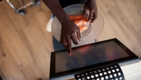 Close-up-shot-from-above:-a-Black-skinned-man-in-a-white-T-shirt-and-black-apron-selects-the-desired-product-on-the-display-of-electronic-scales-and-weighs-vegetables