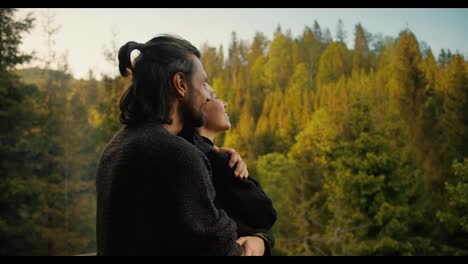 A-brunette-man-approaches-a-girl-on-the-balcony-of-a-country-house,-hugs-her-and-together-they-look-at-the-mountains-covered-with-coniferous-forest