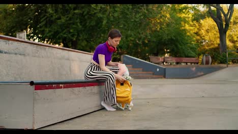 A-young-girl-with-a-short-haircut-in-a-purple-top-takes-out-pink-skates-from-a-yellow-backpack-and-puts-them-on-a-special-area-for-skaters-in-the-park-in-summer