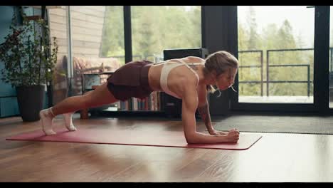 A-blonde-girl-with-tattoos-in-a-white-top-does-a-dynamic-plank-exercise-on-a-special-mat-in-an-industrial-house-overlooking-a-green-forest.-fitness-at-home,-body-and-mind-strengthening
