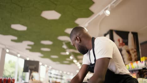 a-Black-skinned-man-in-a-black-apron-lays-out-fruit-and-yakoda-in-special-boxes-on-the-supermarket-counter.-Store-worker-watching-the-products-on-the-counter
