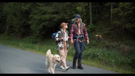 a-guy-and-a-girl-travelers-in-special-clothes-for-hiking-walk-with-their-dog-and-light-coloring-along-the-road-along-the-forest
