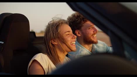 A-bearded-guy-with-curly-hair-in-a-blue-shirt-laughs-with-his-blonde-girlfriend-in-a-convertible-car-on-a-field-background