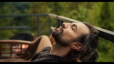 Close-up-shot:-A-young-brunette-man-with-closed-eyes-is-resting-on-a-sofa-on-the-balcony-of-a-country-house-overlooking-the-mountains-and-coniferous-forest.-Spiritual-rest,-outdoor-leisure