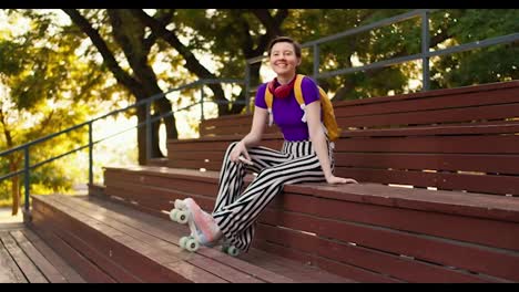 A-girl-with-a-short-haircut-in-a-purple-top,-striped-pants-and-a-yellow-backpack-sits-on-brown-benches-in-the-stands-in-pink-roller-skates-in-a-skatepark-in-summer