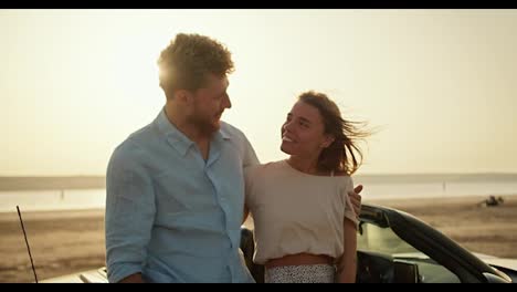 Portrait:-a-bearded-man-in-a-blue-shirt-and-a-blonde-girl-in-a-white-top-are-hugging-and-looking-at-the-camera-on-the-river-bank-during-a-bright-sunset