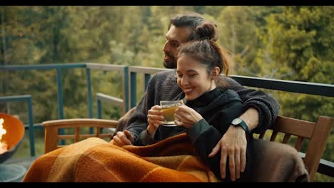The-girl-is-sitting-on-the-couch-next-to-her-brunette-boyfriend-and-drinking-tea,-wrapped-in-a-blanket.-Relax-in-a-country-house-with-a-view-of-the-mountains-and-the-forest