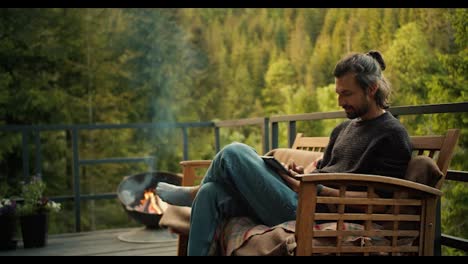 A-man-with-yellow-sits-on-a-sofa-and-works-on-a-tablet-on-the-balcony-of-a-country-house-overlooking-the-mountains-and-coniferous-forest.-Spiritual-break-and-picnic-away-from-the-noisy-city