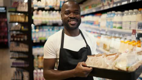 Portrait-of-a-Black-skinned-man-in-a-white-T-shirt-and-black-apron-holding-a-box-of-cottage-cheese,-posing-and-smiling-in-the-dairy-department