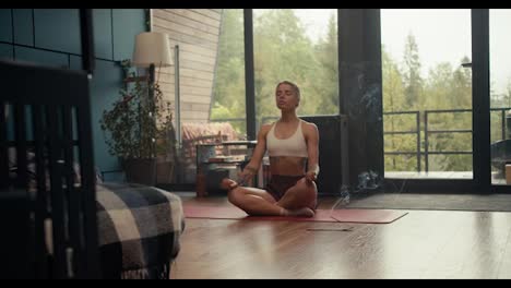 A-blonde-girl-in-a-white-top-with-tattoos-is-meditating-and-using-incense-that-smolders-and-emits-a-small-amount-of-smoke.-Meditation-in-a-country-house-overlooking-a-green-coniferous-forest