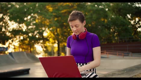 Portrait-of-a-young-short-haired-girl-in-a-purple-swamp-who-sits-on-the-street-in-red-headphones-and-works-on-a-Red-laptop-in-summer