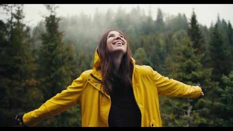A-happy-girl-in-a-yellow-jacket-stands-against-the-background-of-a-coniferous-mountain-forest,-looks-at-the-sky-and-rejoices-in-a-little-rain