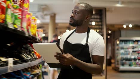 a-Black-skinned-man-in-a-white-t-shirt-and-a-black-apron-walks-along-the-windows-of-a-supermarket-and-takes-inventory-using-paper-and-a-tablet