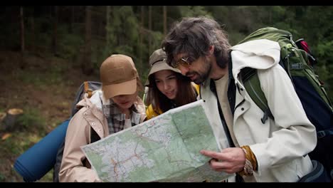 A-trio-of-hikers-in-trekking-attire-with-large-backpacks-survey-their-path-on-a-large-paper-map-they-hold-in-their-hands.-Orientation-in-the-area-during-the-travell