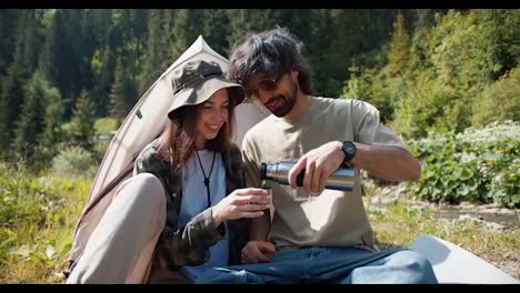 A-brunette-guy-in-yellow-sunglasses-pours-tea-from-a-thermos-to-a-brunette-girl-in-a-cap,-they-sit-near-the-tent-against-the-backdrop-of-a-green-forest