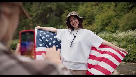 A-blonde-girl-adjusts-and-says-How-to-stand-up-a-brunette-girl-in-a-white-t-shirt-with-an-American-flag-for-a-better-photo-against-the-backdrop-of-the-forest