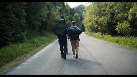 Rear-view-of-a-guy-and-a-girl-tourists-in-special-clothes-for-hiking-with-large-backpacks-walk-along-the-road-along-the-green-forest