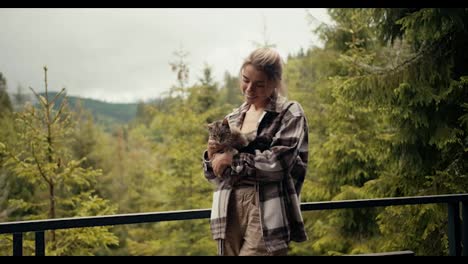 Happy-blonde-girl-petting-her-cat-on-the-balcony-of-a-country-house-overlooking-a-coniferous-forest-in-the-mountains
