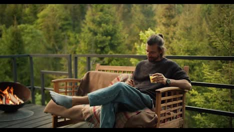 A-man-with-a-yellow-drink-in-his-hand-sits-on-a-sofa-and-works-on-a-tablet-on-the-balcony-of-a-country-house-overlooking-the-mountains-and-coniferous-forest
