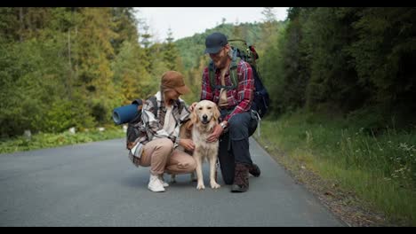 A-guy-and-a-girl-tourists-in-special-clothes-for-hiking-stand-near-the-road-and-stroke-their-light-colored-dog.-A-couple-of-blondes-against-the-backdrop-of-a-green-mountain-forest