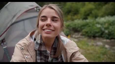 Portrait-of-a-happy-girl-blonde-tourist-who-sits-near-the-tent-on-the-background-of-the-forest
