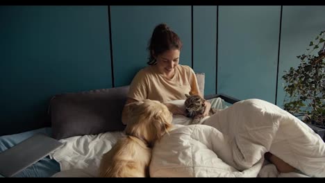 A-brunette-girl-lies-on-a-bed-near-her-dog-and-cat.-The-cat-does-not-like-the-presence-of-the-dog,-but-the-girl-reconciles-them.-Interact-pets-at-home