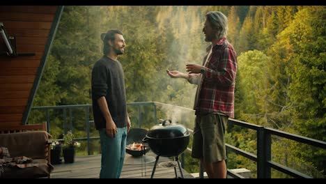 A-couple-of-friends-chat-while-cooking-on-the-grill.-A-brunette-guy-in-a-green-sweater-lifts-the-lid-of-the-grill-and-thick-white-smoke-comes-out.-Picnic-with-a-view-of-the-mountains-and-coniferous-forest