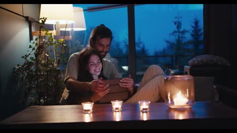 Romantic-evening:-a-guy-and-a-girl-are-sitting-on-a-devan-in-a-room-lit-by-candles-and-looking-at-the-tablet-screen.-Room-overlooking-the-evening-coniferous-forest