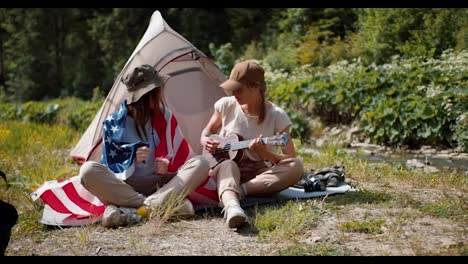 2-American-girls-are-sitting-with-the-USA-flag-while-camping-on-the-lawn-near-the-forest