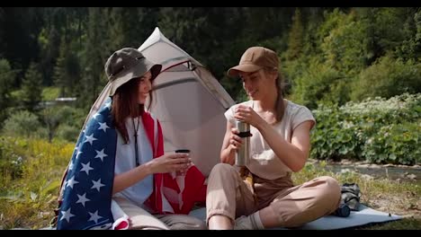 A-blonde-girl-in-a-cap-pours-tea-for-a-brunette-girl-who-is-wrapped-in-the-flag-of-the-United-States-of-America-near-the-tent-against-the-backdrop-of-a-green-forest