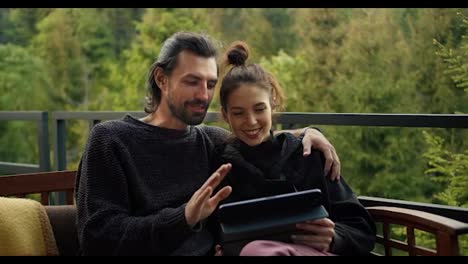 A-brunette-guy-and-a-girl-are-sitting-on-a-devan-and-looking-at-a-tablet-screen-against-the-backdrop-of-mountains-and-forests.-Rest-in-a-country-house