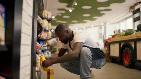 a-Black-skinned-man-in-a-white-t-shirt-and-black-apron-crouched-near-the-display-case-in-order-to-arrange-the-goods-on-the-shelf.-Tracking-the-positions-of-goods-on-the-counter,-inventory