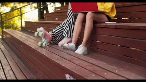 Close-up-shot:-a-girl-in-striped-pants-and-roller-skates-and-a-girl-in-shorts-and-white-sneakers-sit-on-a-brown-bench-and-work-on-a-red-laptop-in-a-park-in-summer