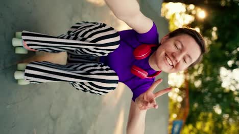 Vertical-video:-Happy-girl-with-a-short-haircut-in-a-purple-top-and-red-headphones-in-striped-pants-rides-sitting-on-pink-roller-skates-in-a-skate-park-in-summer