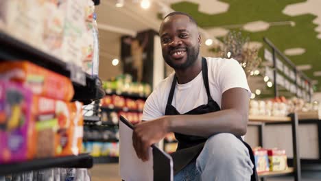Portrait-of-a-Black-skinned-man-in-a-white-t-shirt-and-black-apron-crouched-near-the-shop-window,-looks-at-the-camera-and-smiles
