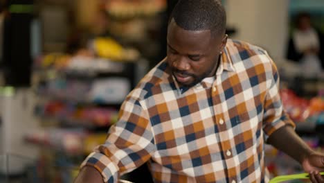 Overview-of-a-Black-man-in-a-plaid-shirt-walks-along-the-counter-in-the-supermarket-and-takes-the-berries-in-the-box-that-he-liked
