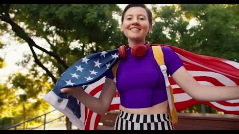 Portrait-of-a-young-girl-in-a-purple-top-and-red-headphones,-who-wraps-herself-with-the-US-flag-in-the-park-in-summer.-A-young-patriot-girl-looks-at-the-camera-and-smiles