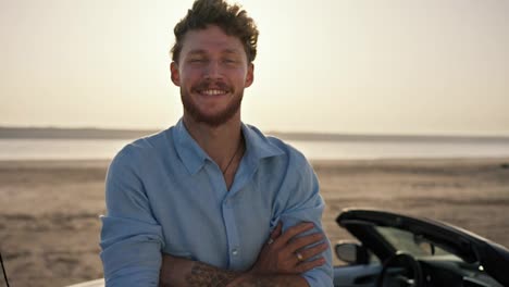 Portrait:-A-bearded-man-with-curly-hair-wearing-a-blue-shirt-is-crossing-his-arms,-smiling-and-standing-next-to-a-white-convertible-against-the-yellow-sky-on-the-beach