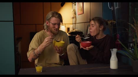 Happy-couple-girl-and-guy-stoles-sit-in-the-kitchen-in-the-morning-and-eat-cereal-with-milk-in-a-cozy-room