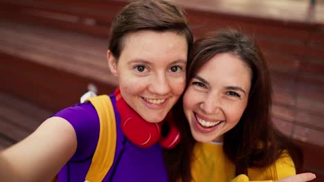 Portrait-of-a-brunette-girl-in-a-yellow-sweater-and-a-girl-with-a-short-haircut-in-a-purple-top-and-red-headphones-who-take-a-selfie-on-a-brown-bench-in-a-skatepark-in-summer