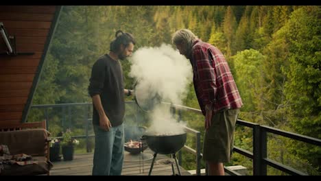 Two-guys-check-the-food-they-cook-on-the-grill-and-blow-high-fives-to-each-other,-because-they-are-doing-great.-Thick-white-smoke-comes-from-the-brazier-against-the-backdrop-of-mountains-and-coniferous-forests